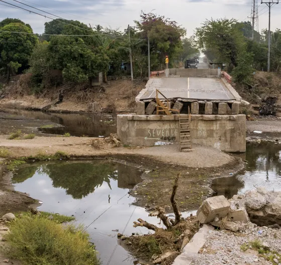 A two-lane bridge is shorn in half across a patchy river on an overcast day