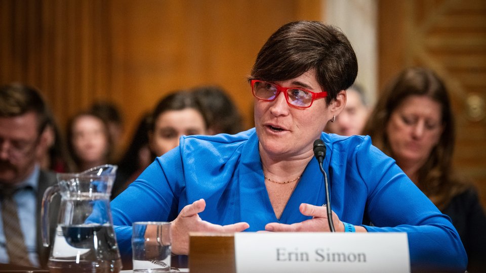 Woman in blue shirt and red glasses sitting at desk with microphone
