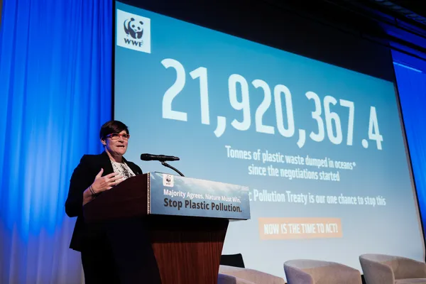 WWF's Erin Simon stands at a lectern in front of a screen that shows how much plastic has been dumped into the ocean since plastic pollution negotiations began.