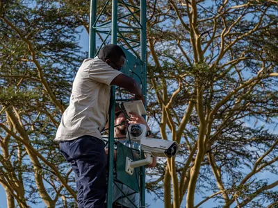 close-up of a man in a green tower setting up a FLIR nightvision camera against a backdrop of trees