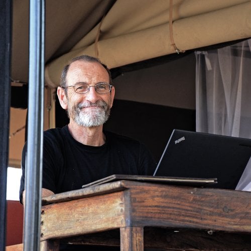 An older white man with a salt and pepper beard and gray hair sits at a desk with his computer and smiles gently.