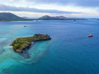 A chain of islands surrounded by barrier reefs in blue water with a red boat anchored offshore