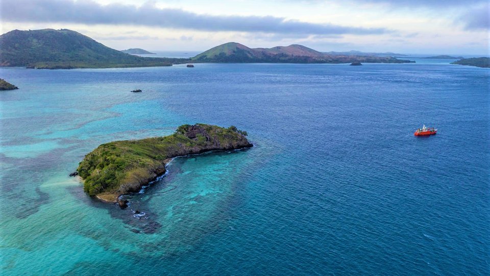 A chain of islands surrounded by barrier reefs in blue water with a red boat anchored offshore