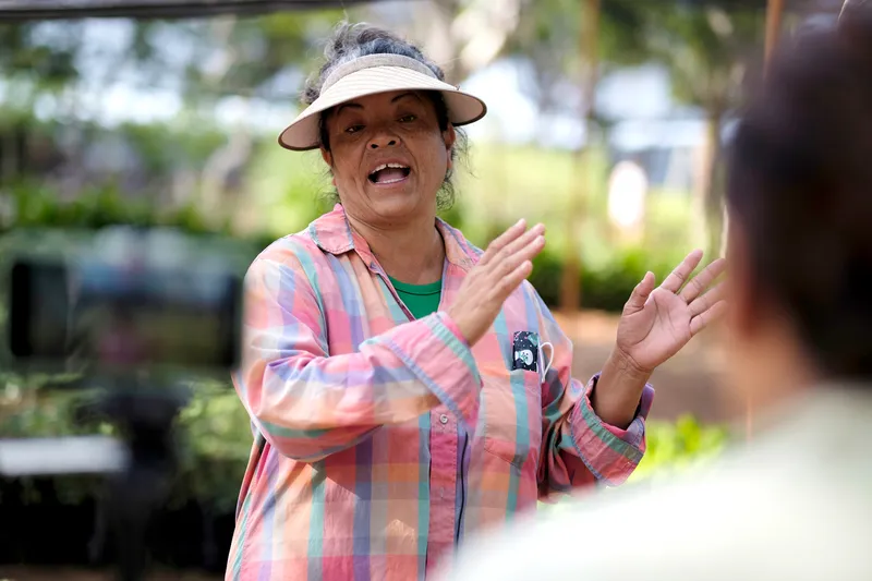 A woman in a pink plaid shirt and a wide-brimmed visor talking with her hands centered and raised.