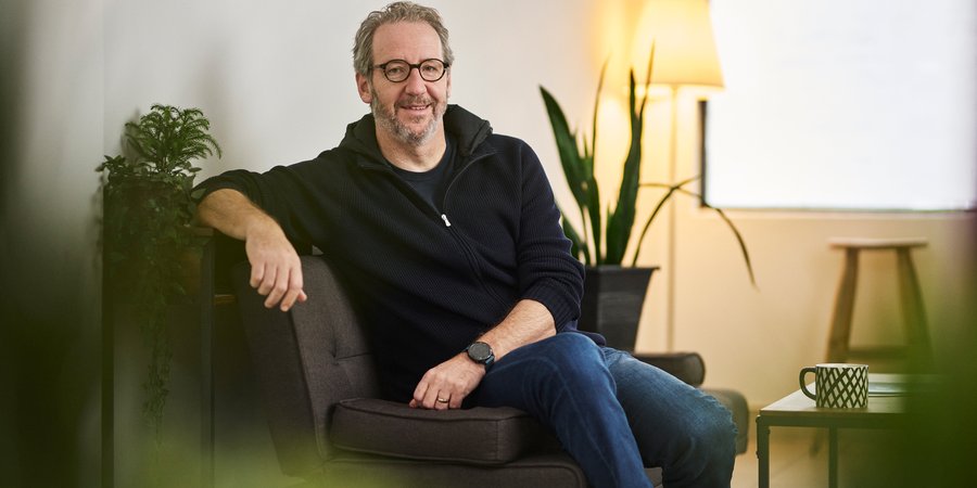 A man sittiung on a green chair in a modern indoor setting with a plant, a coffee table and a cup of coffee