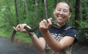 A young woman holds an eel in her hands. She is smiling and looking at it. She is wearing round glasses and has her brown hair in a bun.