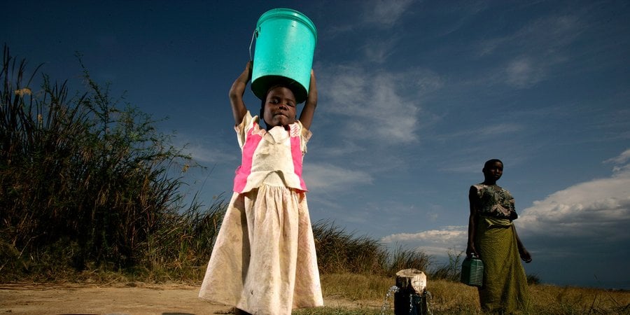 A girl in a white dress holds a green bucket above her head and smiles at the camera