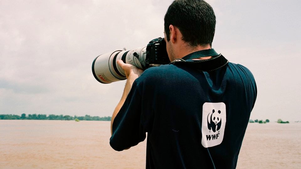 A photographer wearing a long-sleeve WWF t-shirt points a camera to take a photo of the vast Greater Mekong River.