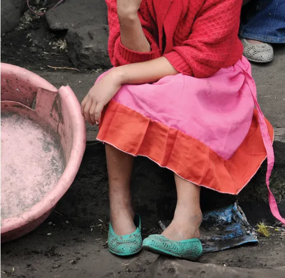 The bottom half of a woman sitting on a step wearing a pink and orange sweater and dress and turquoise shoes next to an empty plastic bin