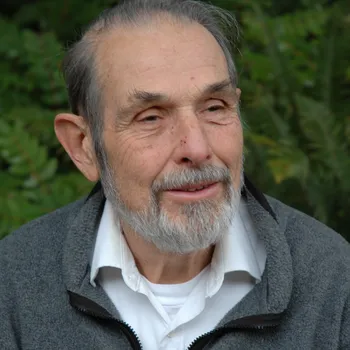 An older man with a salt and pepper beard wears a gray zip jacket and white button down.