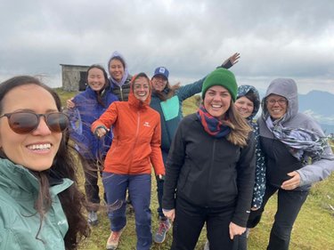 An all woman team members pose for group photo on top of a windy mountain.