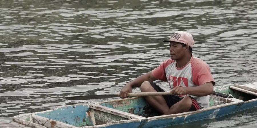 A man in a tshirt and pink baseball cap sits inside a skinny blue canoe on the water