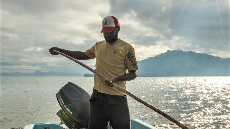 A man in a yellow tshirt and baseball cap stands in a small blue fishing boat holding a long paddle out at sea with the sun shining and a mountain range behind him