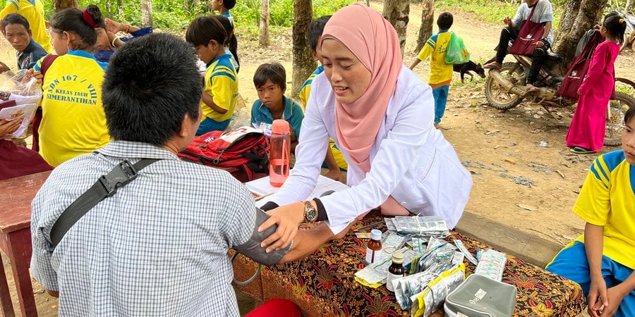 Herni reaches across a table to take someone's blood pressure