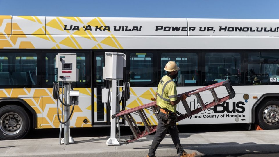 A worker carrying a ladder walks past an electric bus and charging station.