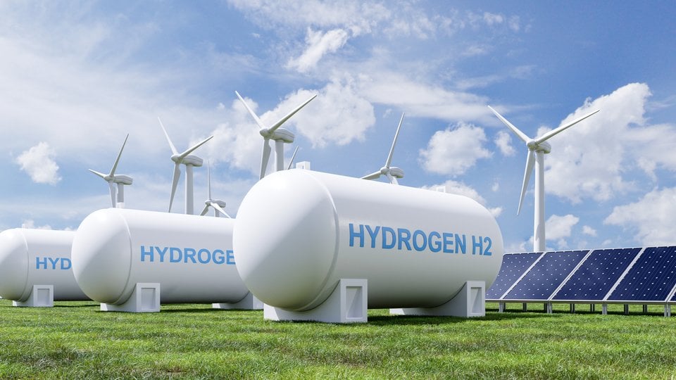Three white hydrogen tanks in the foreground with wind turbines and solar panels in the background against a blue and white sky