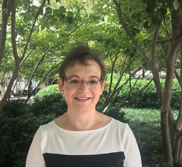 A photo of a smiling woman with short hair and glasses, with branches and green leaves in the background.