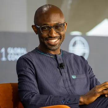 A bald Black man wearing black rimmed glasses sits and smiles in an orange armchair in front of a conference panel screen.