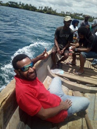 A man wearing sunglasses and a red shirt smiles at the camera while sitting in a small wooden boat on the ocean