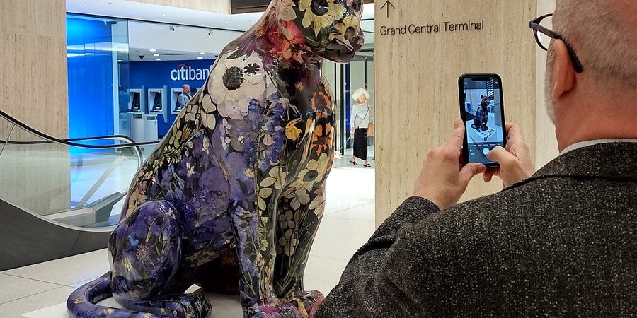 A man taking a photo with his iPhone of a decorated jaguar statue in an indoor venue with an escalator in the background.