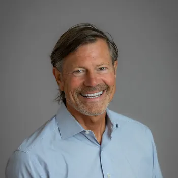 Smiling headshot of a white man with semi-long hair tucked behind his ear who wears a blue button down.