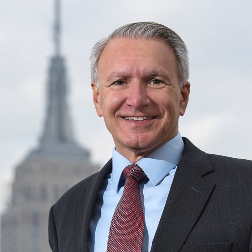 Joe Proto smiles at the camera, wearing a suit with a tall building seen behind him