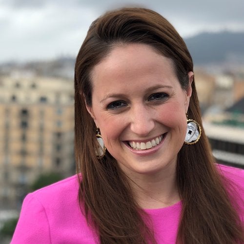 Julia Cardozo smiles at the camera, wearing a bright pink top and a cityscape visible behind her