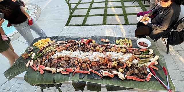 People gather around a large table covered with grilled banana leaves and food