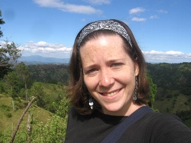 A white woman with medium length brown hair smiles at the camera with green mountains and a blue sky in the distance.