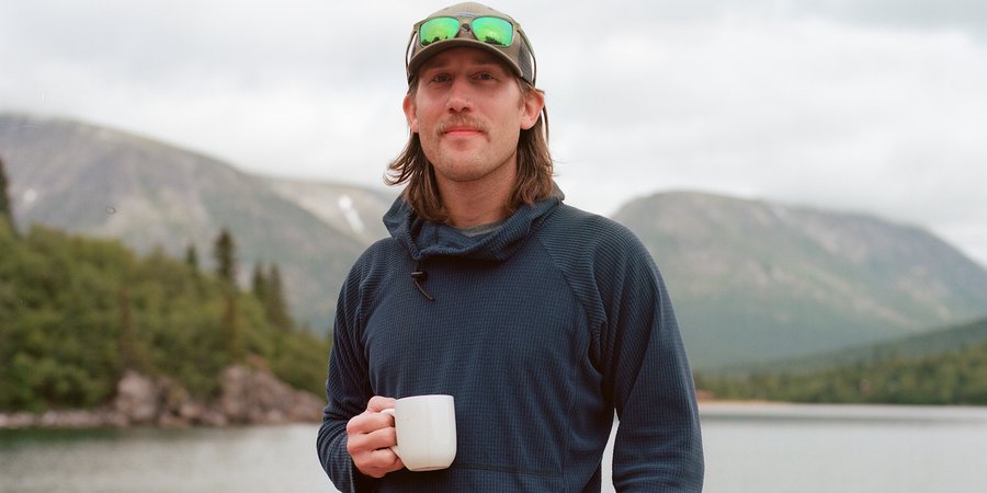 A white man with long blonde hair and mustache holds a white coffee mug and has one hand in his pocket. He is wearing outdoor gear and is standing in front of a vast landscape with water and mountains. He is wearing a hat and sunglasses are propped on top