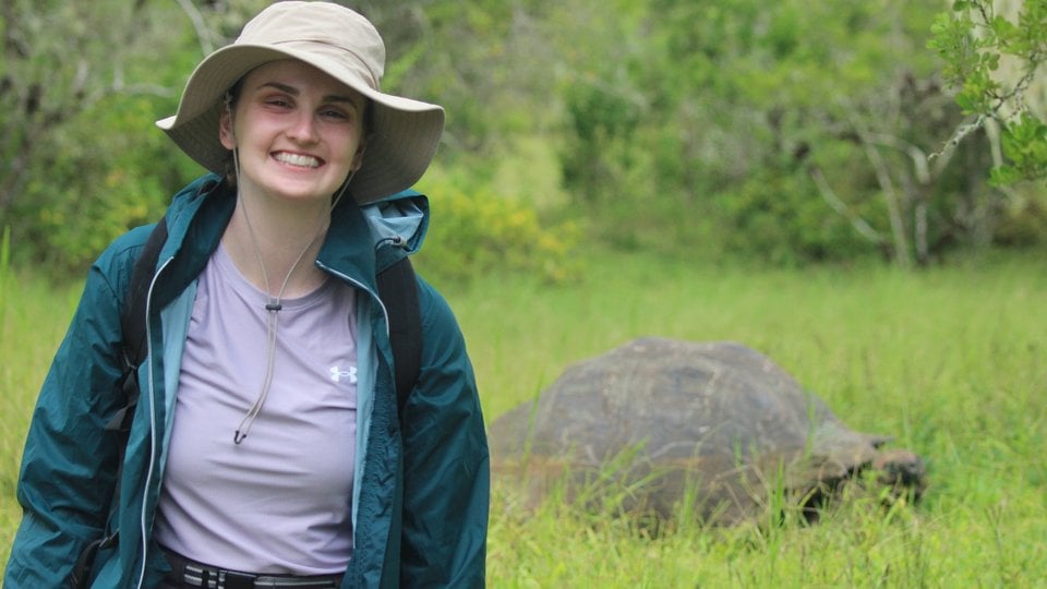 A woman in a hat and outdoor gear in the foreground with a large tortoise turtle in the green grass in the background.