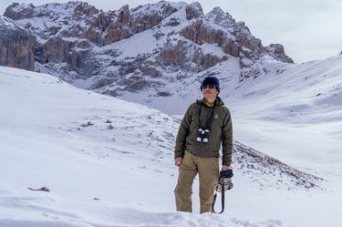 A Tibetan man stands in snow with binoculars and his camera