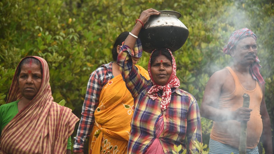 Mahua holds a large pot over her head as she walks in front of several people