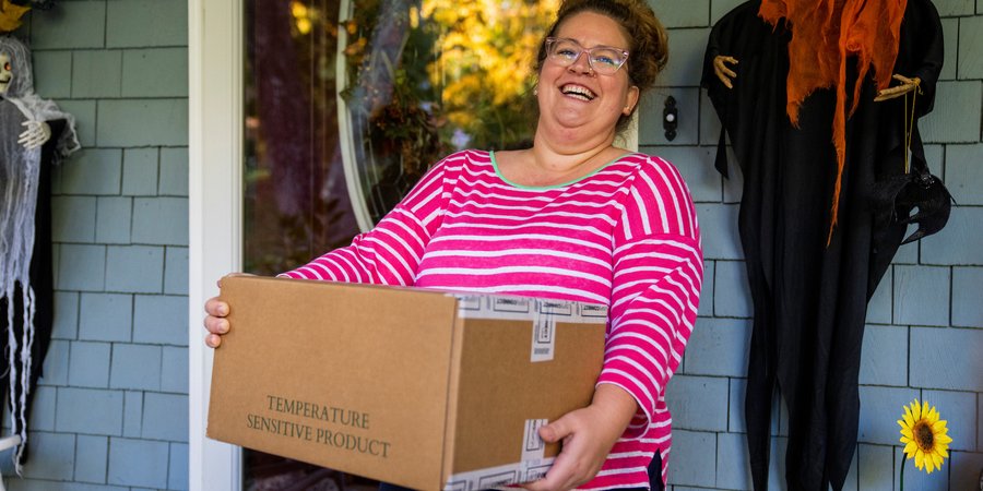 A woman in a pink shirt smiles on a porch holding a box of vegetables to mail