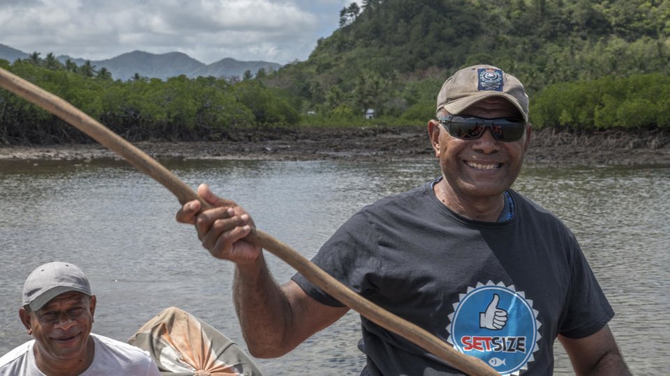 A photo showing two men int he foreground with a paddle, a body of water and trees are in the background.