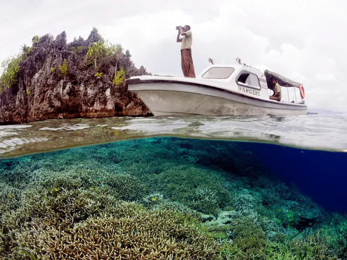 A person stands with binoculars on the front of a boat amid a coral reef.
