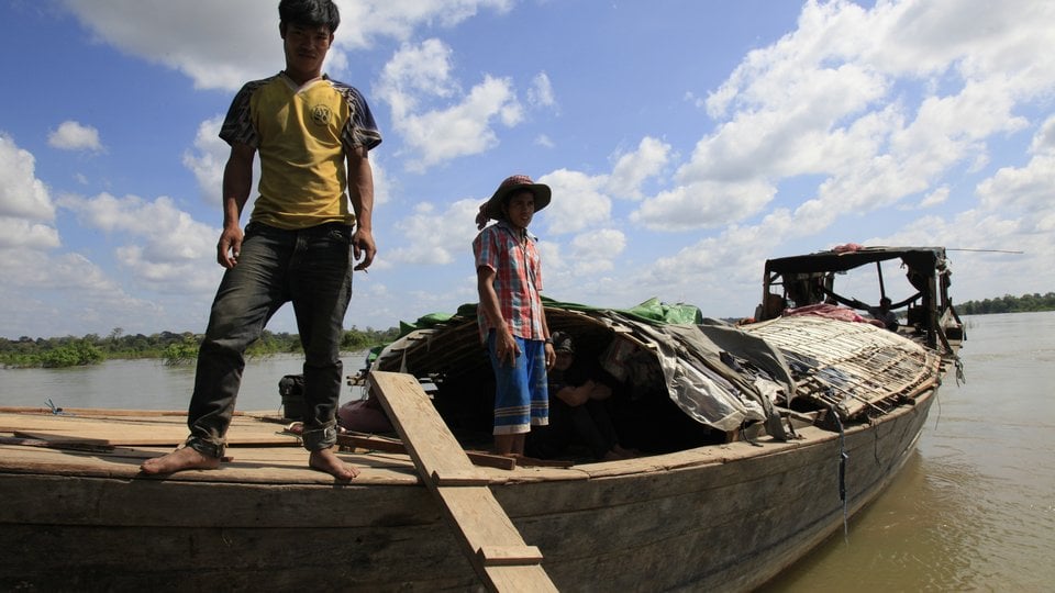 Two people on a wooden boat on a flat murky river with a cloudy sky in the background.