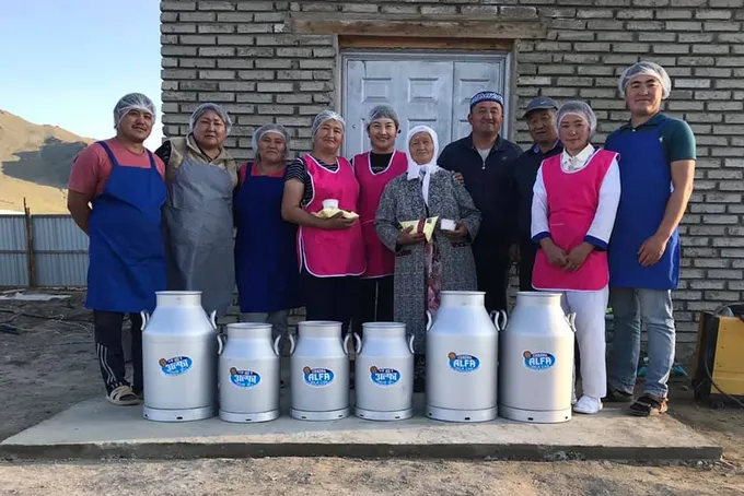 A group of people wearing blue or pink aprons and hair nets stand in a row smiling at the camera behind a row of metal milk jugs
