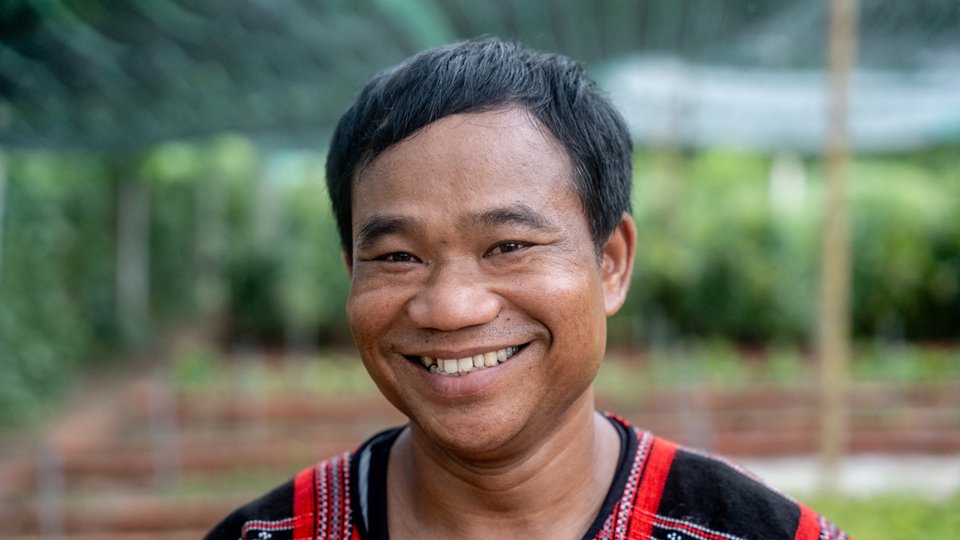 A closeup color photo portrait of a man in the foreground with a village in Vietnam in the blurred green background