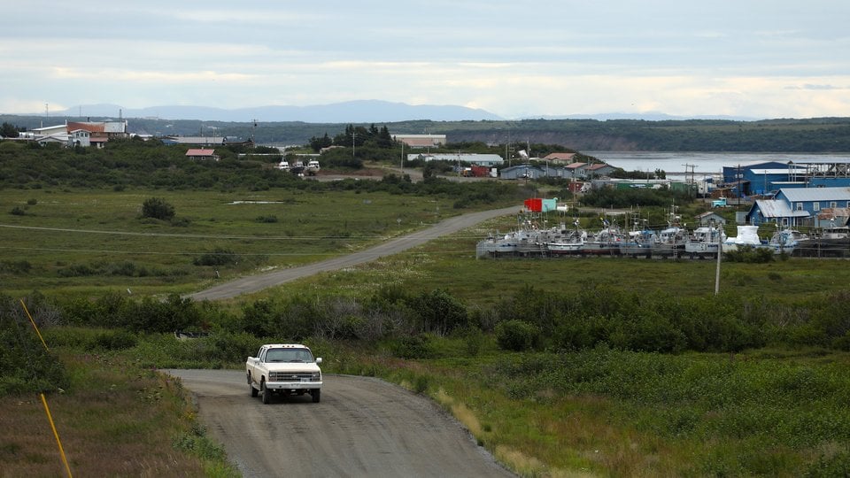 A car drives up a road with the village of Naknek in the background