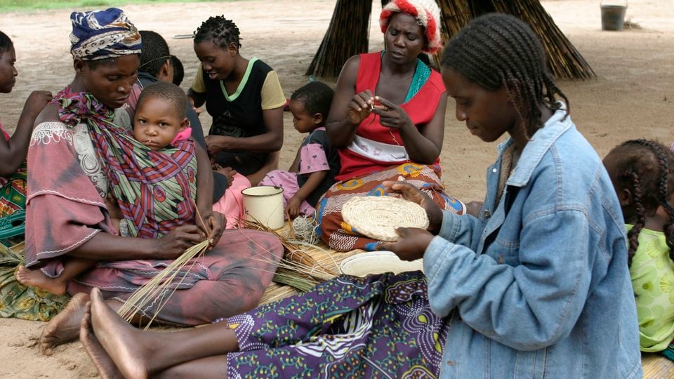 A group of women and children sitting on the ground outside working on handcrafts.