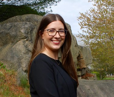 A woman with long brown hair and glasses smiles at the camera in front of large rocks.