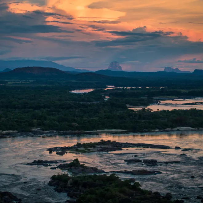 An orange and blue cloudy sky at sunset over a dark river.