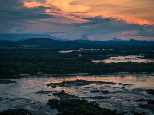 An orange and blue cloudy sky at sunset over a dark river.