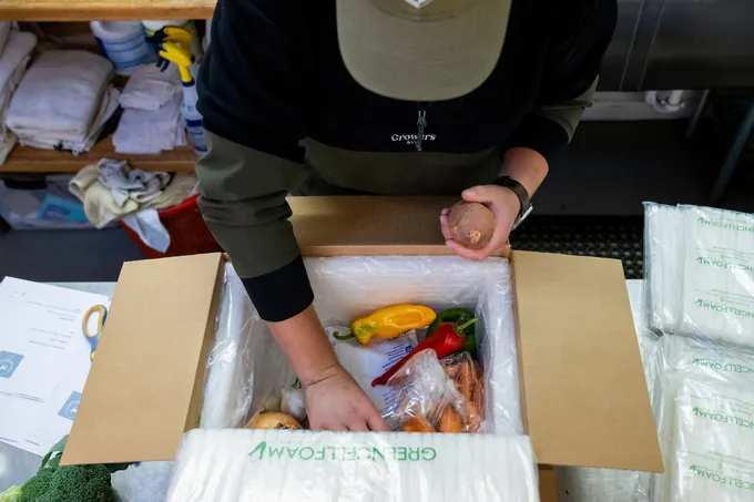 A person leans over a box of vegetables that they are packing to mail