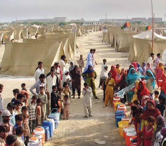 A crowd of people created an aisle using colorful buckets on dirt with a road with tents on either side in the background