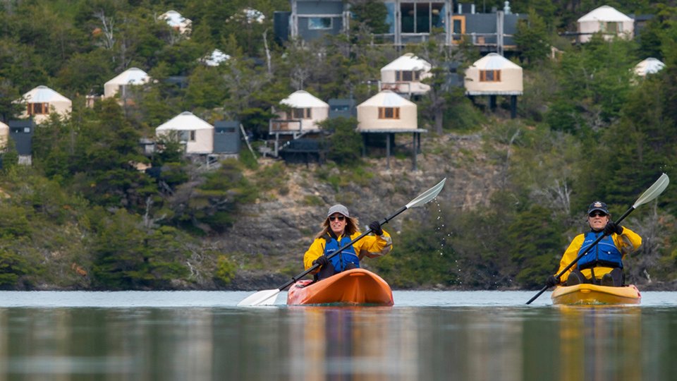 Two kayakers paddle towards the camera with round tents on platforms dot the slope behind them.