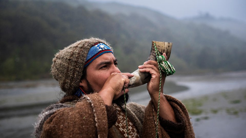 A color photo showing a man with a pipe in the foreground with mountains and a waterway in the misty background