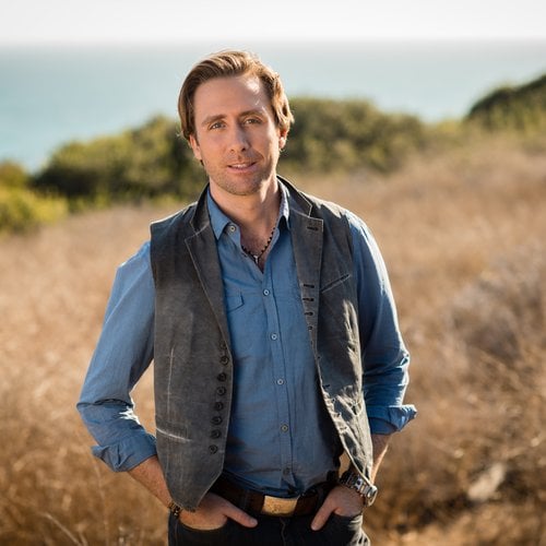Philippe Cousteau wearing a vest and a blue shirt with a body of water in the background.