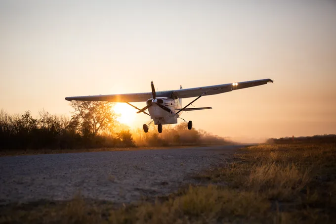A color photo showing the silhouette of a small airplanen landing against the backdrop of a sunset on a tree-lined runway
